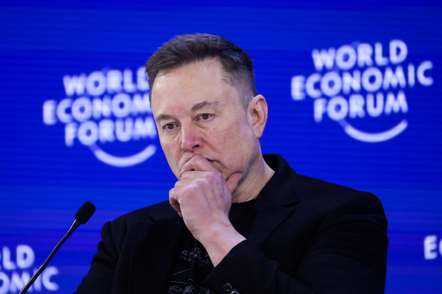 Elon Musk, in front of a blue "World Economic Forum" background, puts his hand to his mouth.