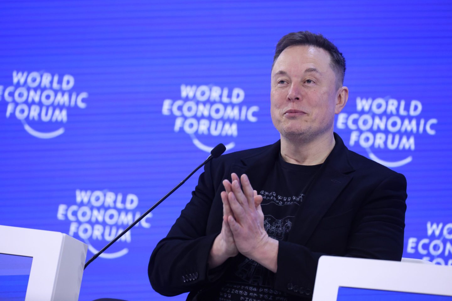 Elon Musk, wearing all black and in front of a blue background, presses his hands together.